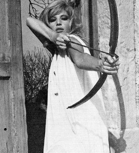 Black and white photo of a woman in a long white dress aiming a bow and arrow, standing outdoors against a rustic wooden door and stone wall.